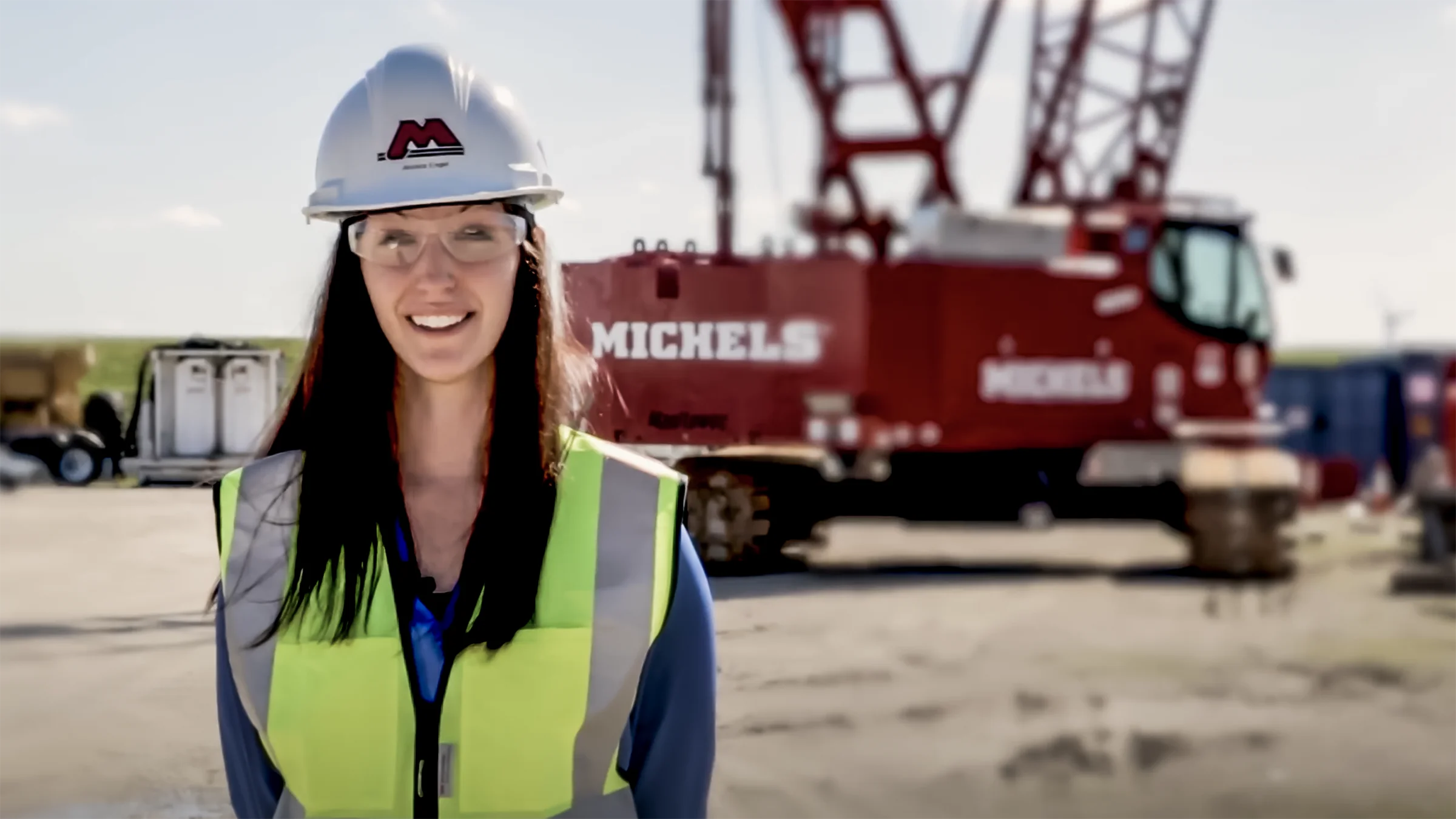 A woman wearing safety equipment stands near an excavator