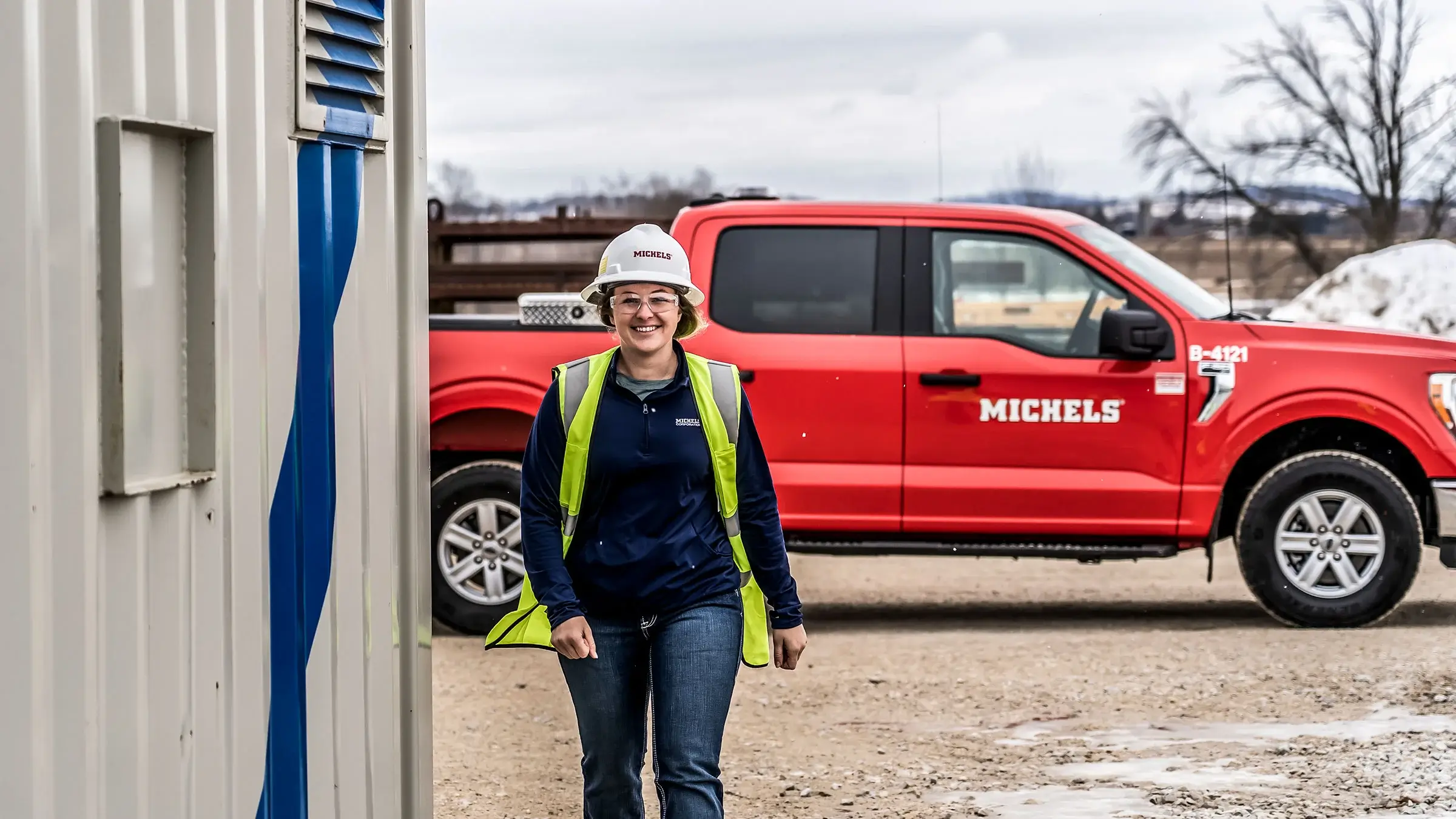 A woman walks by a red Michels pickup truck in a yard.