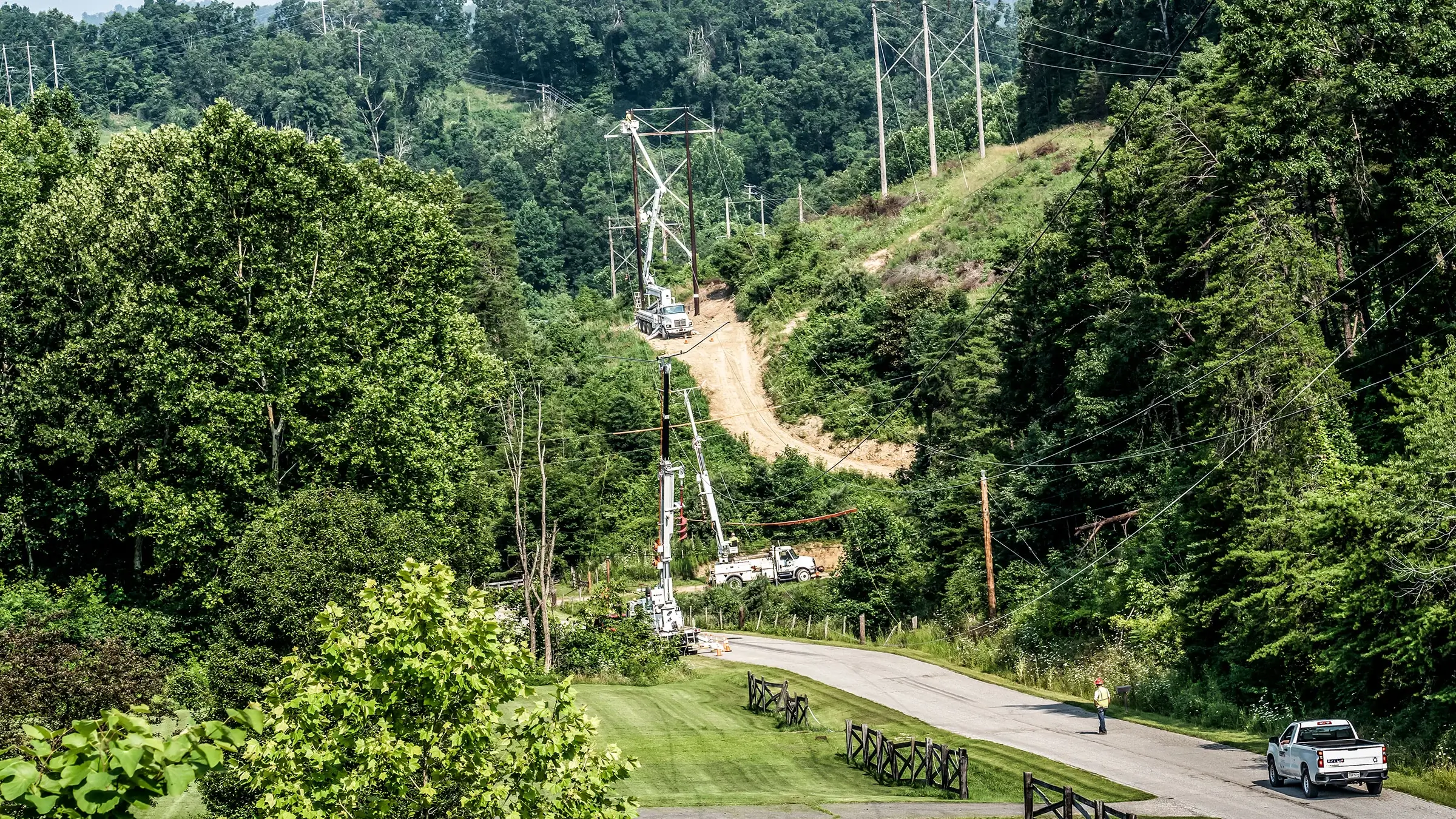 A power crew consisting of several bucket trucks operates on transmission lines in a wooded area.