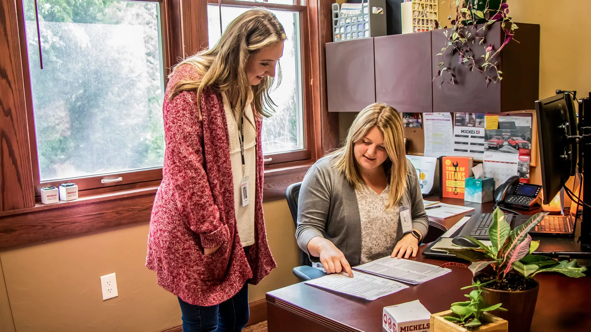 Two women stand near an office desk as one points to a paper.