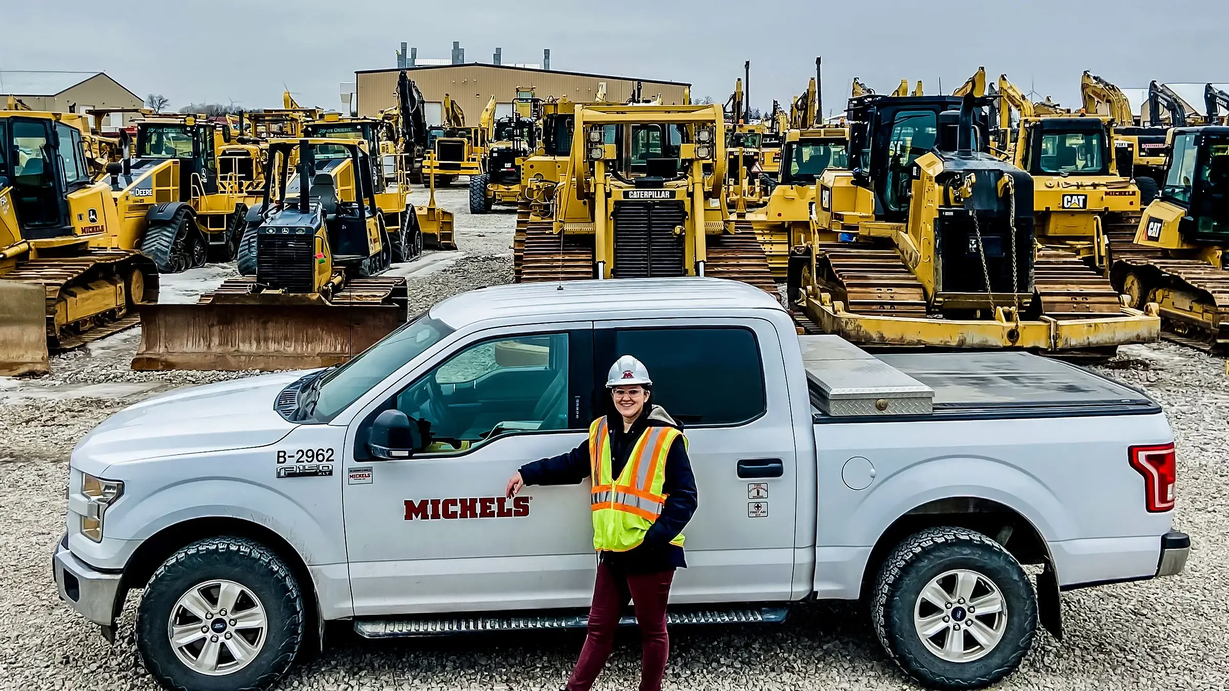 A woman stands in next to a white Michels pickup in a yard full of equipment.