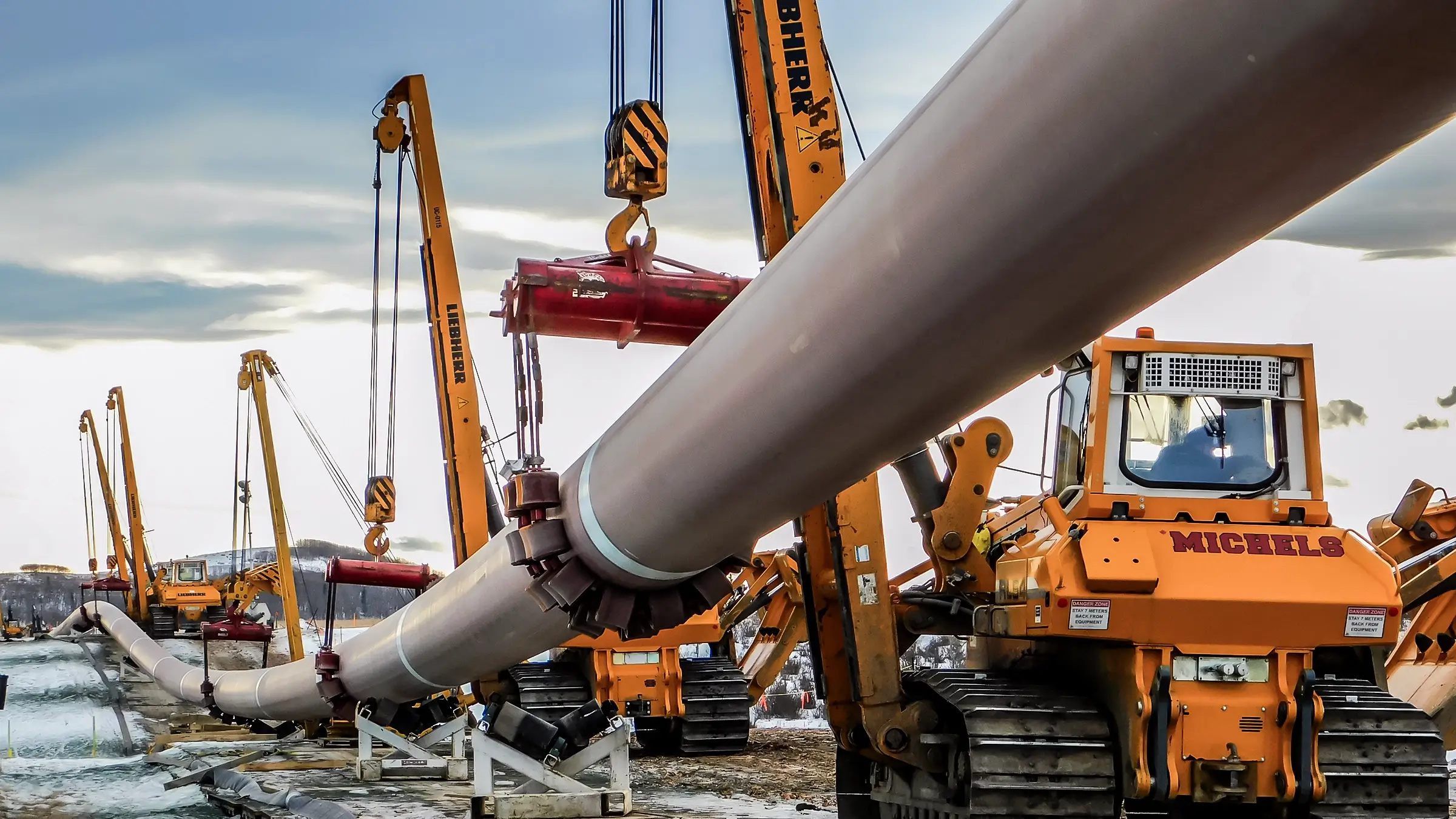 Several pipelayers carry a large diameter pipe through a snowy environment.