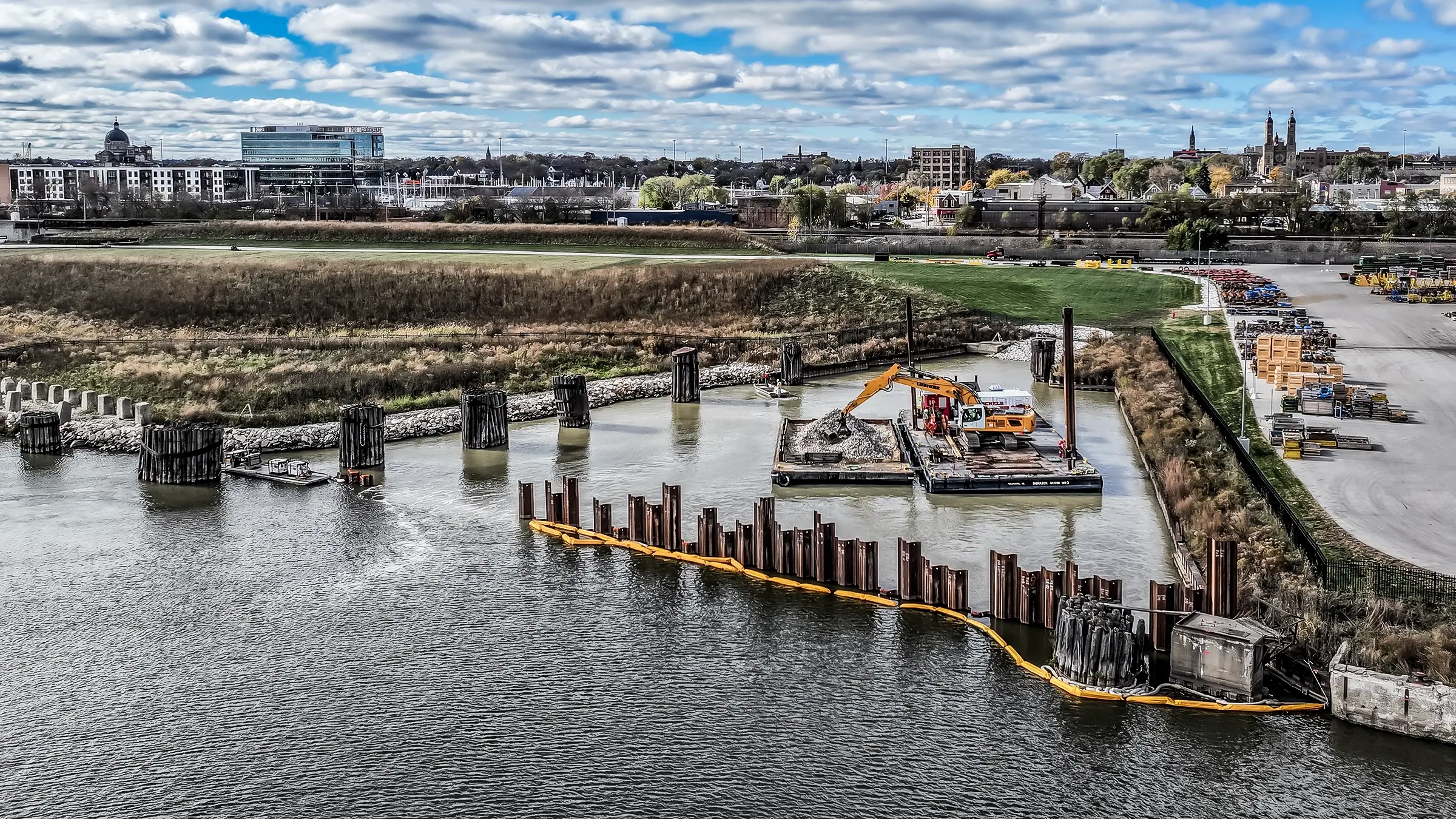 An excavator handing aggregates work on a barge offshore near a city.